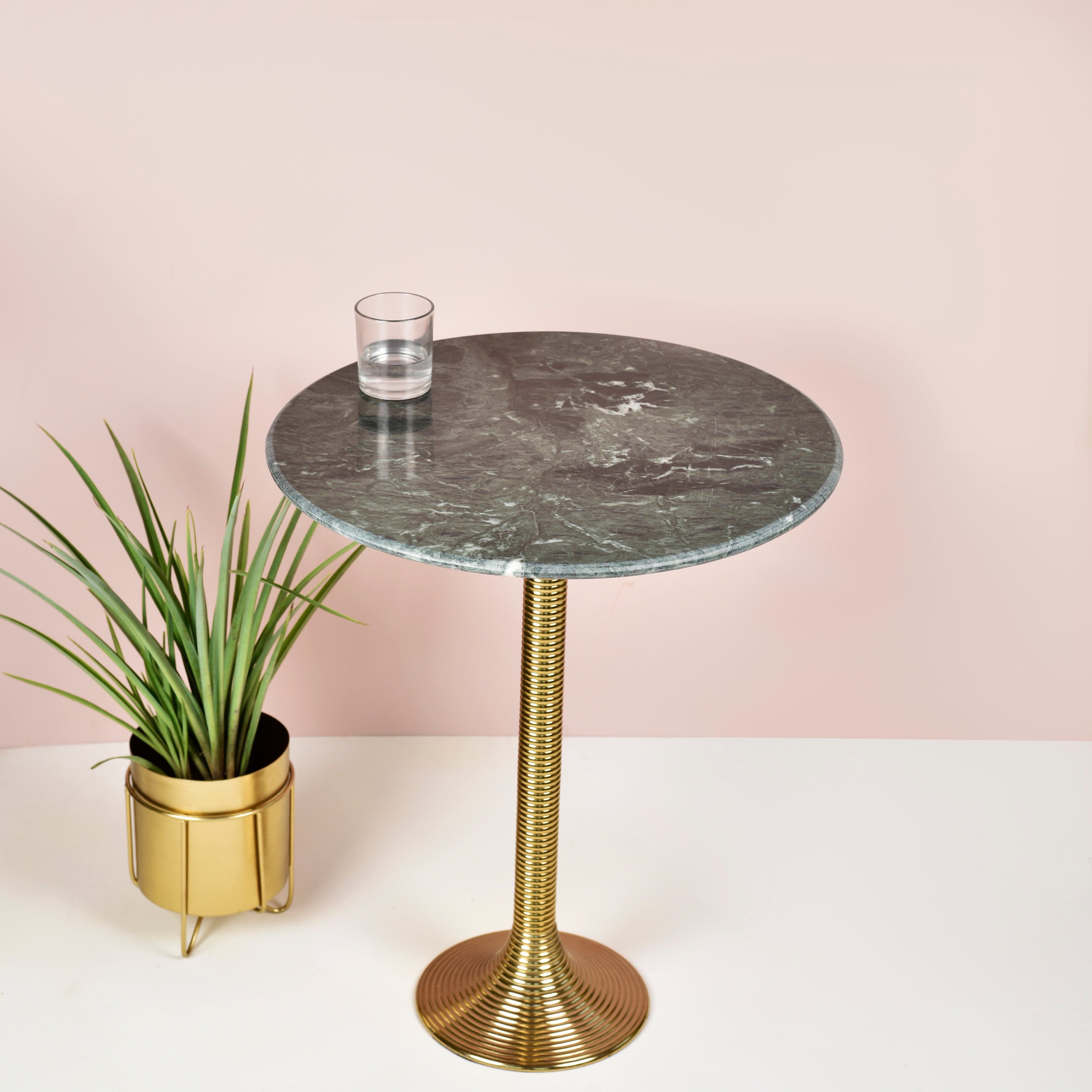 An elegant ribbed metal coffee table with a round white marble top and a gold-colored, circular base, set against a pink background with a potted plant to the left.