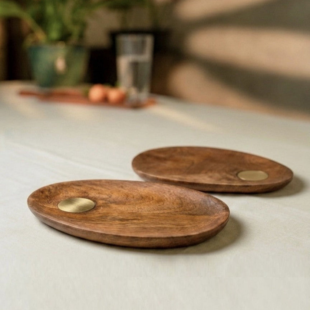 Two wooden coasters with brass centers on a light surface, with a blurred background of plants and a glass.