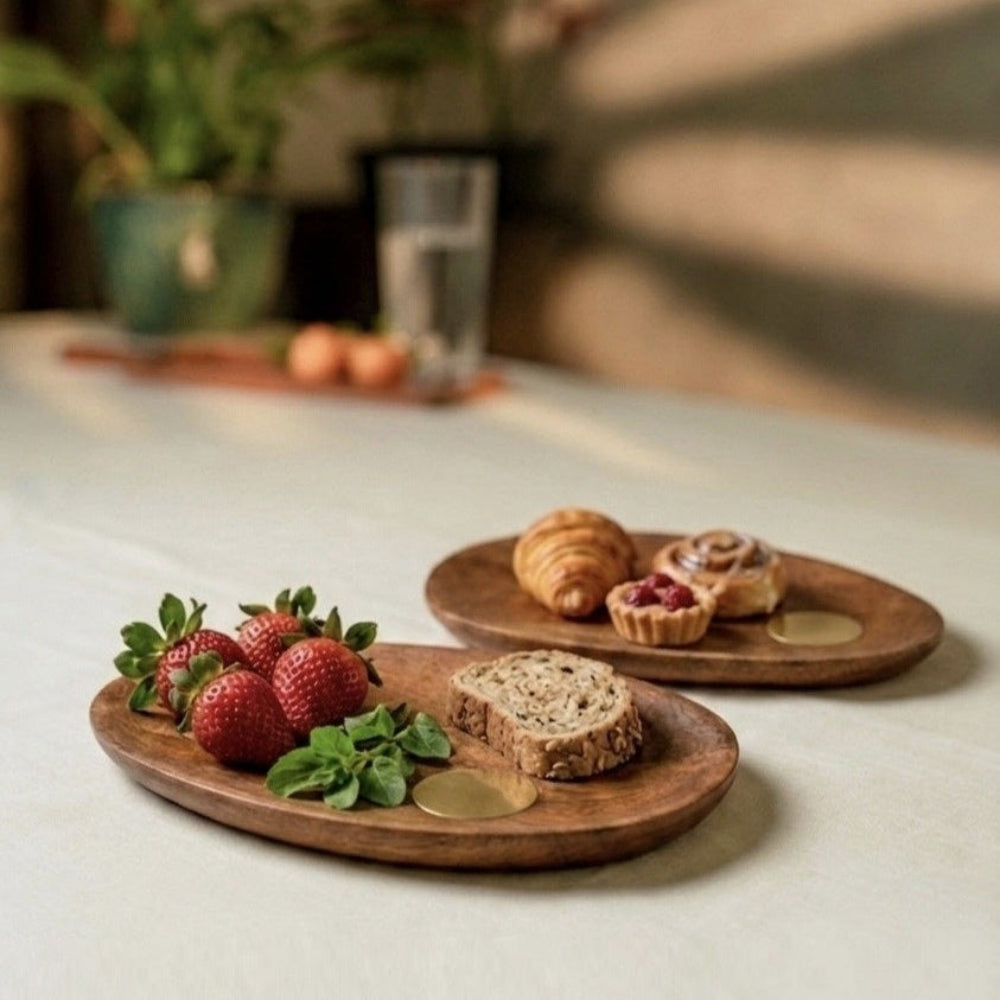 Two wooden plates with food on a light surface, with plants and a glass in the background.