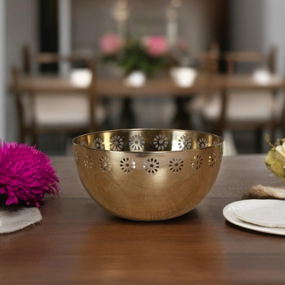 A gold metal bowl with a floral pattern, placed on a wooden table with decorative items around it.