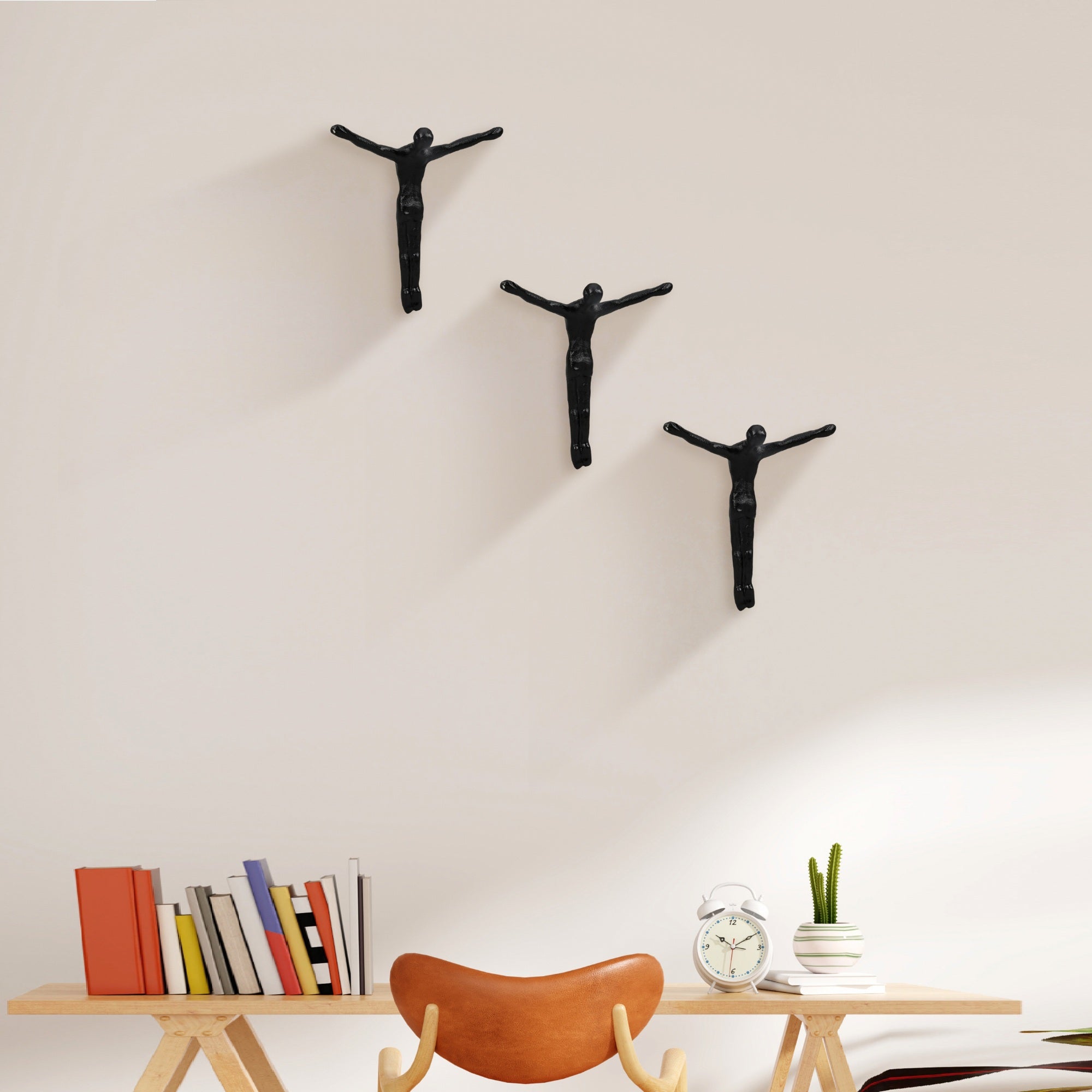 Three black wall-mounted figures on a light beige wall with a wooden table and books below.