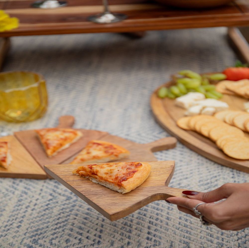 A set of five pizza slice-shaped wooden platters, with one slice lifted to show the wood texture, displayed on a table with a pizza and other food items.