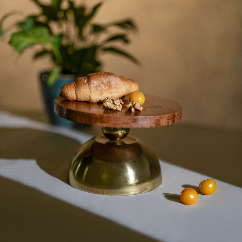 Wooden and brass cake stand with a croissant and small fruits on a light surface.