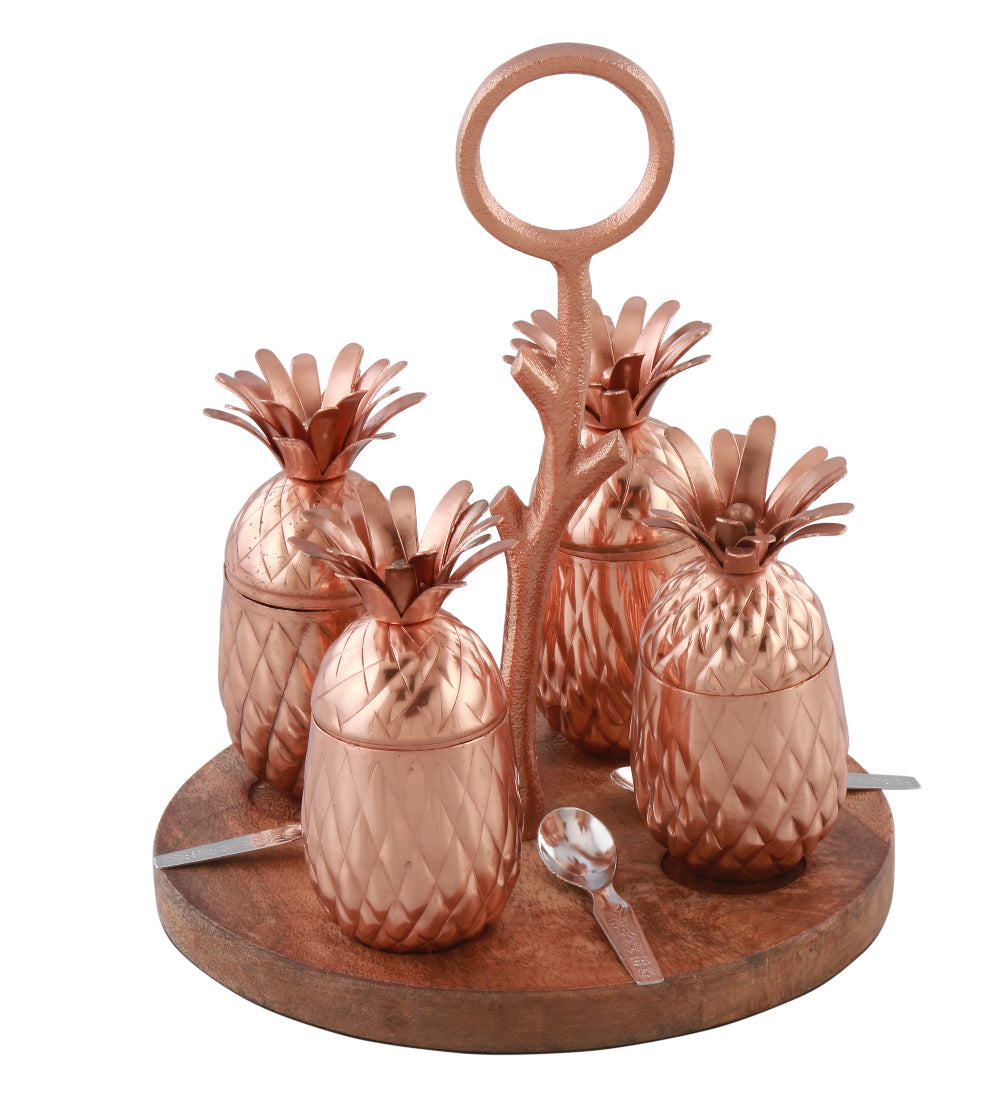 A set of copper-finished pineapple-shaped jars with a wooden base tray and metal handles, displayed with decorative pineapples and spoons on a table.
