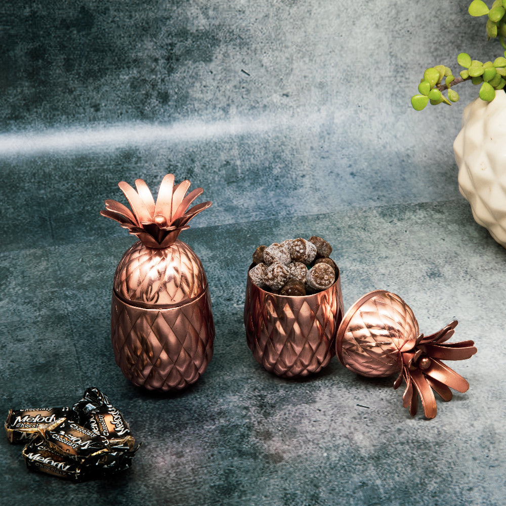 A set of copper-finished pineapple-shaped jars with a wooden base tray and metal handles, displayed with decorative pineapples and spoons on a table.