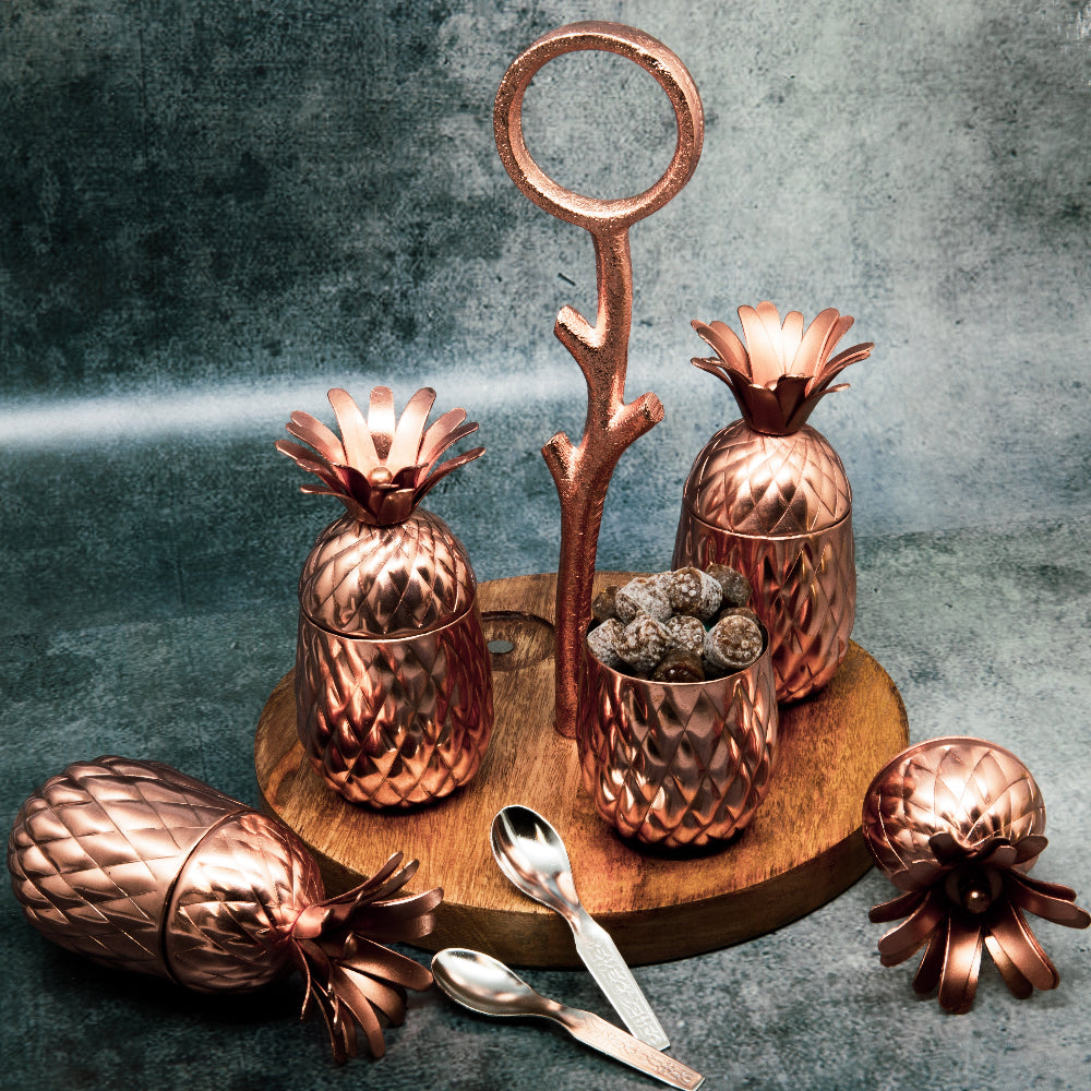 A set of copper-finished pineapple-shaped jars with a wooden base tray and metal handles, displayed with decorative pineapples and spoons on a table.